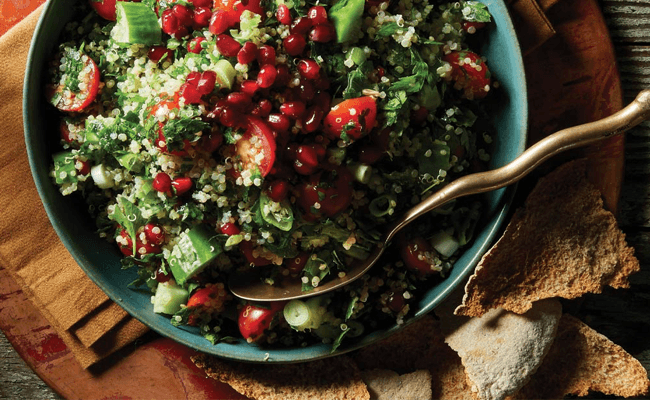 Quinoa Tabbouleh with Pomegranate Seeds in a blue dish with a gold metal spoon and a side of crackers