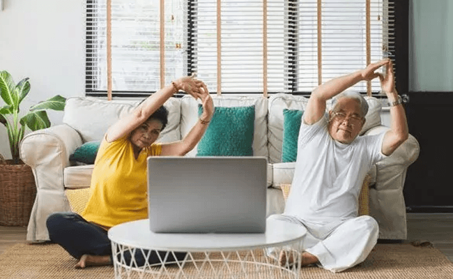 Two older Asian people sitting on the floor in front of a laptop stretching to an online qigong video