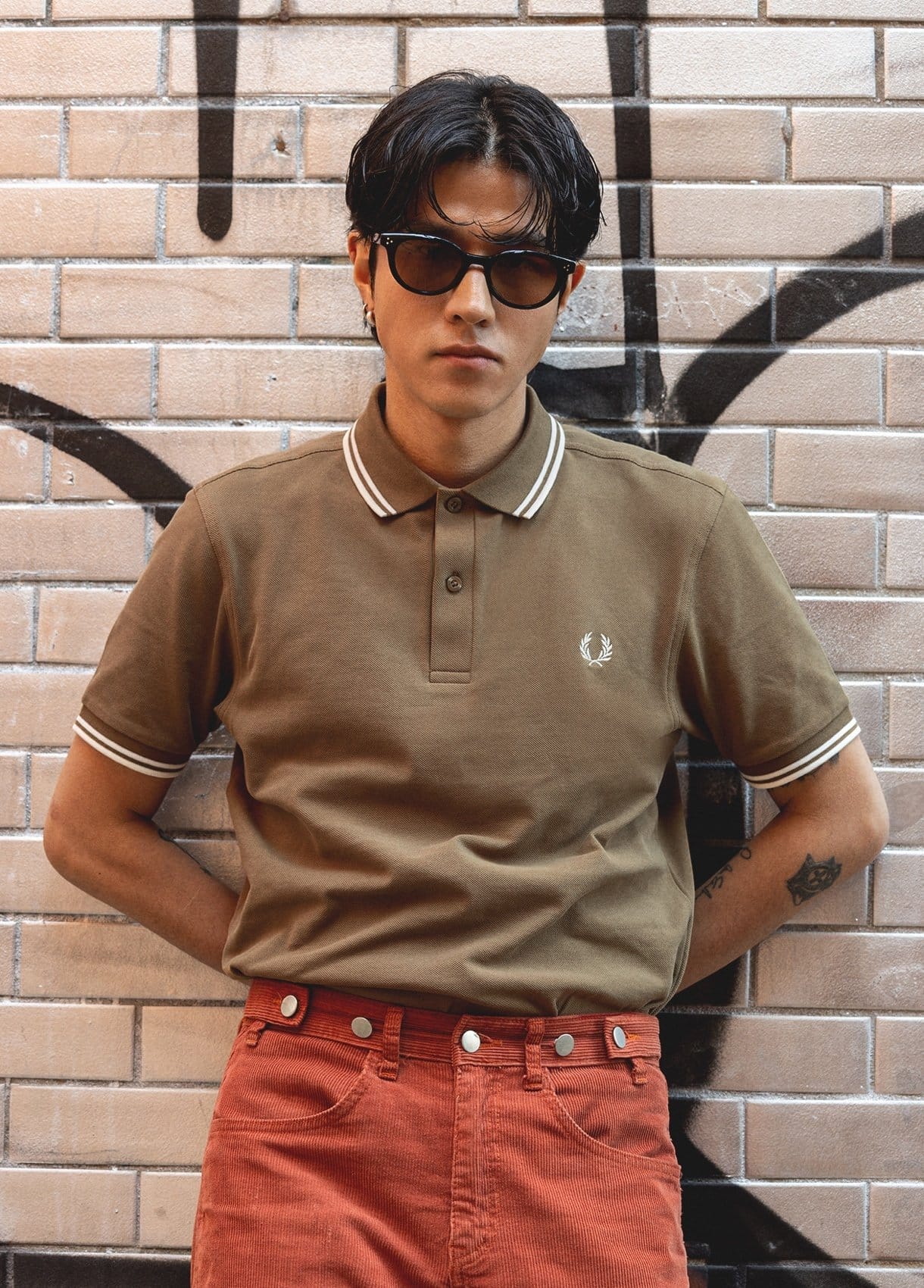 Male model leaning against a wall wearing a brown fred perry polo shirt