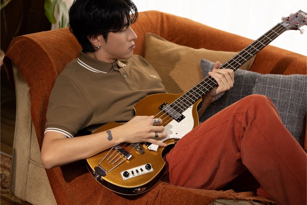 Male model reclined on sofa wearing a brown fred perry polo shirt playing an electric guitar