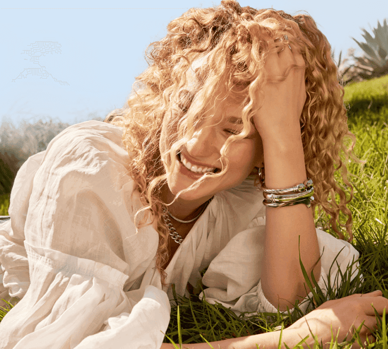 A blond woman laying down on the grass on a sunny day, adorned with silver necklaces & bracelets