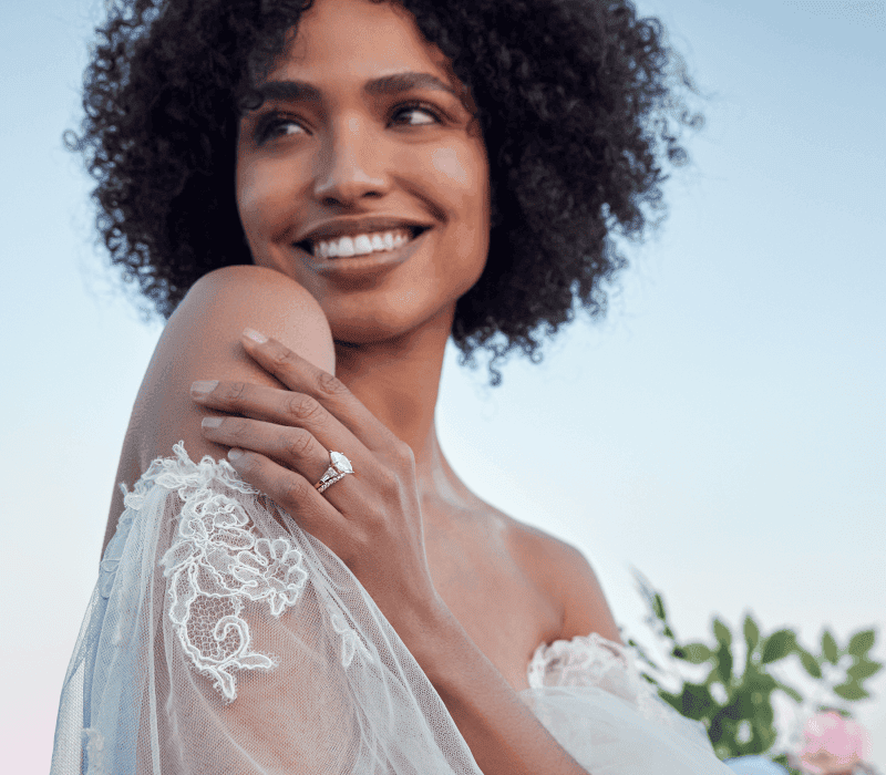 Smiling bride with curly hair showcasing elegant diamond engagement ring on left hand.