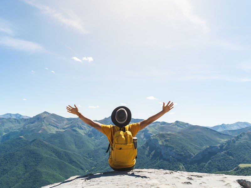 A young man with hands raised in triumph, sitting atop a mountain while wearing a vibrant yellow backpack, embracing the spirit of adventure and achievement.