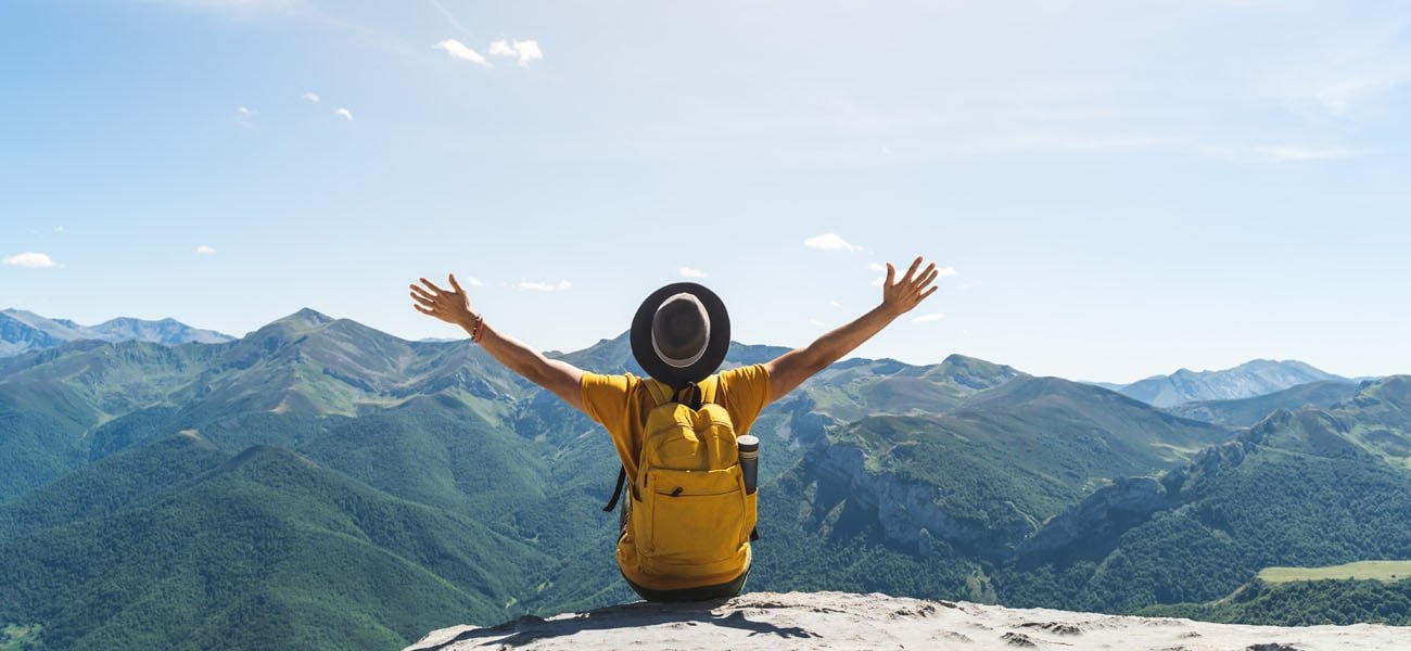 A young man with hands raised in triumph, sitting atop a mountain while wearing a vibrant yellow backpack, embracing the spirit of adventure and achievement.