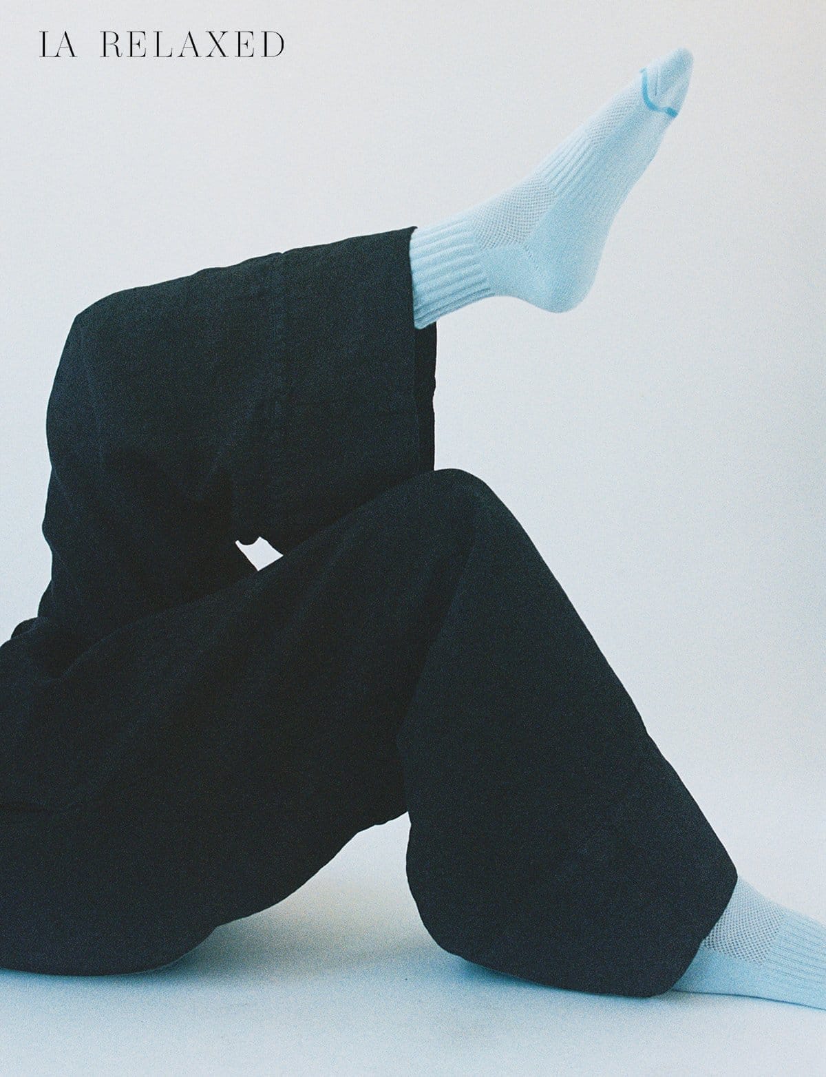 Cropped image of model wearing baggy black linen pants with blue socks and legs crossed.