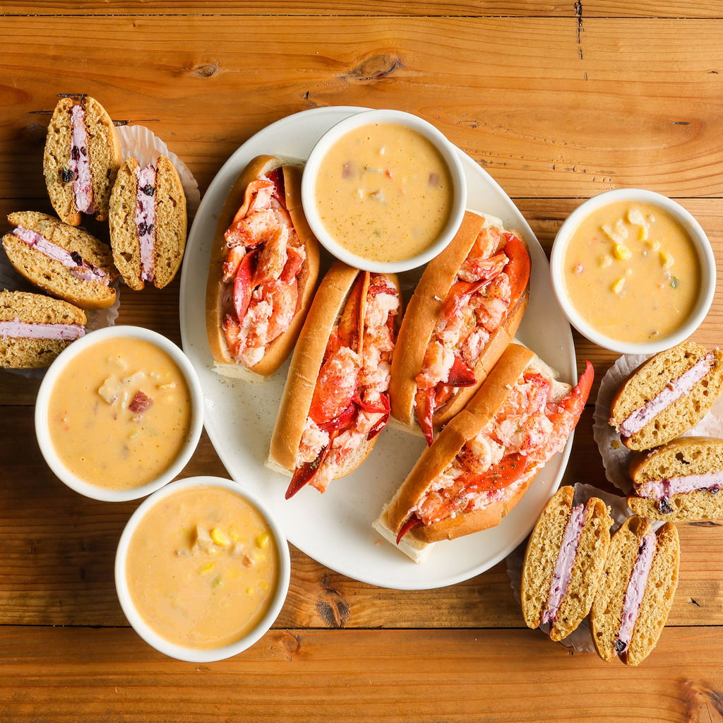 A wooden table with a plate of four lobster rolls, four cups of chowder, and four whoopie pies cut in half