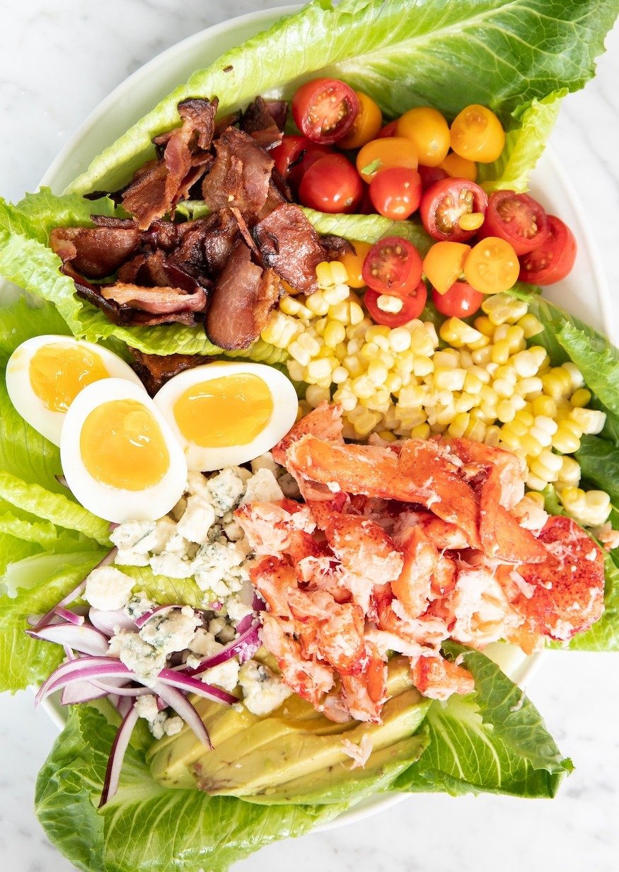 A salad topped with lobster meat in a white plate on a white marble countertop