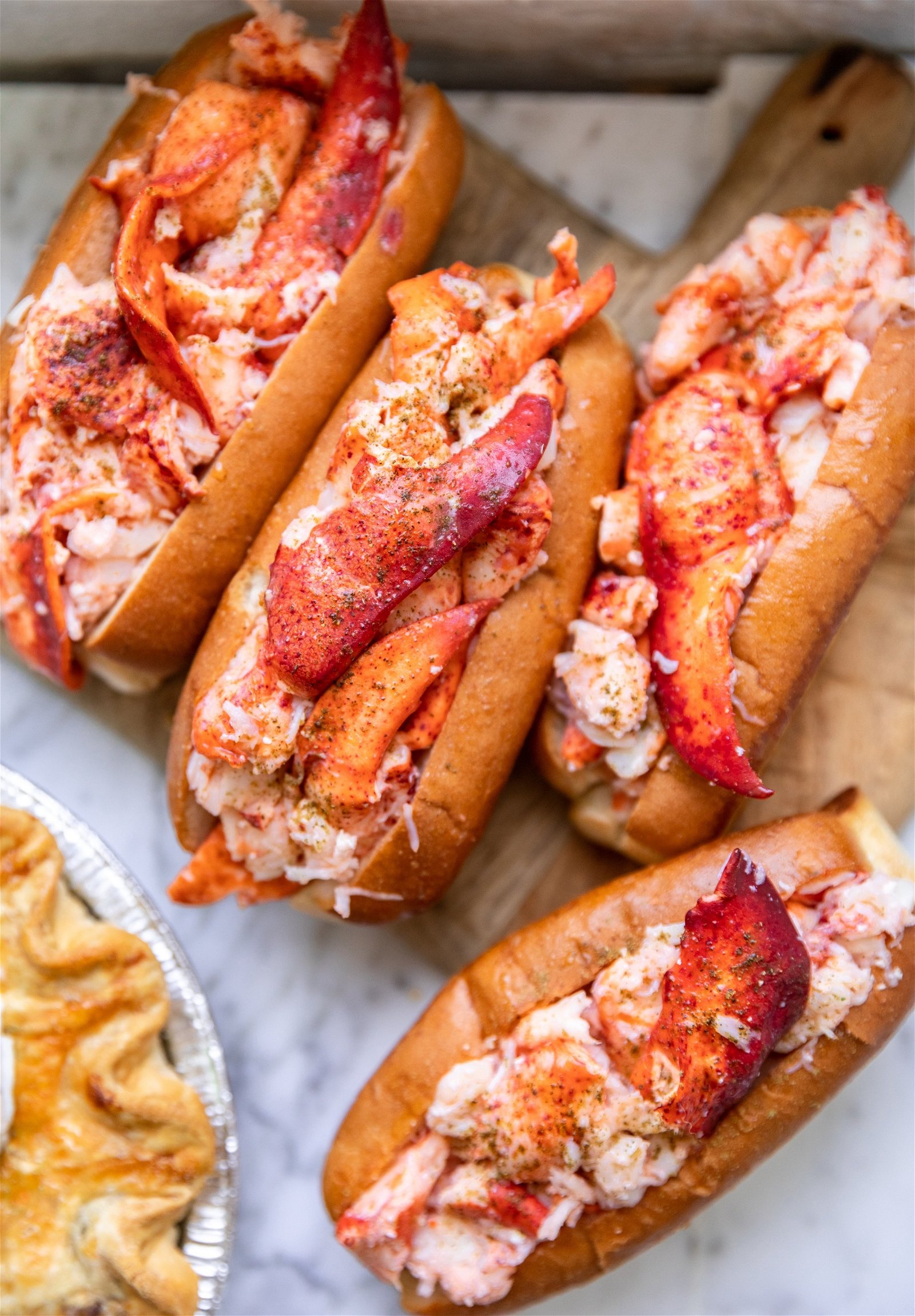 Four lobster rolls on a wooden cutting board next to a pie