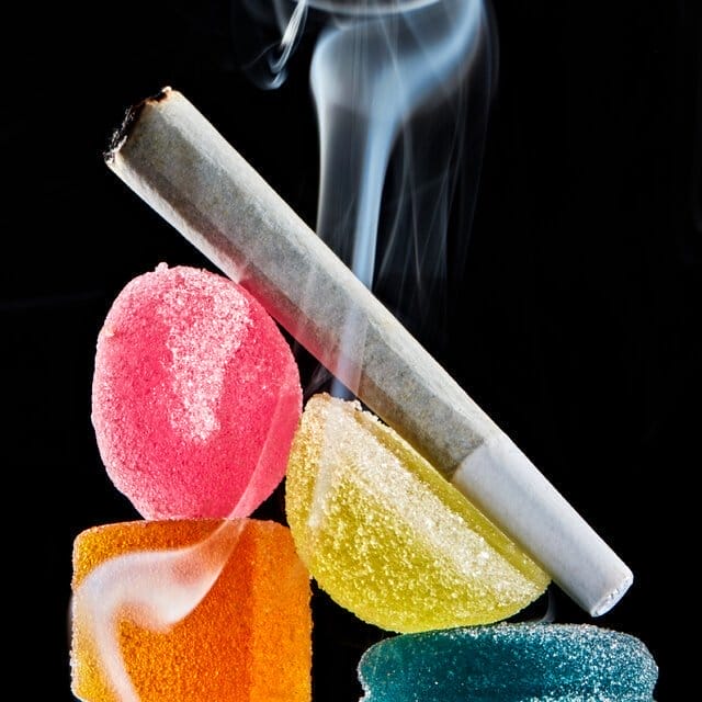 A still life of a smoking joint stacked on top of edibles against a black backdrop.