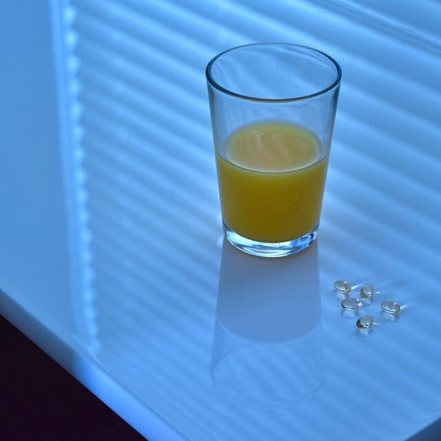 A photograph of a half-full glass of orange juice next to five gel supplements on a shiny white table. The blinds are closed and the room is dark.
