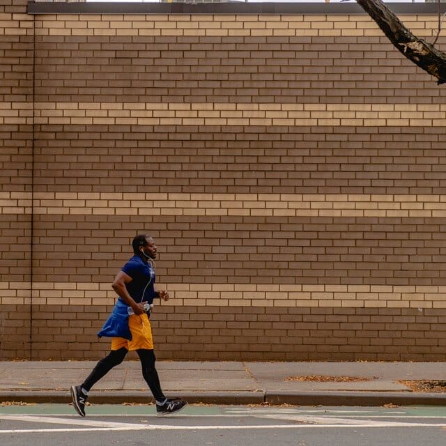 A man runs beside a striped brick wall in orange shorts and a blue top.