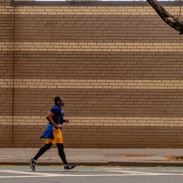 A man runs beside a striped brick wall in orange shorts and a blue top.