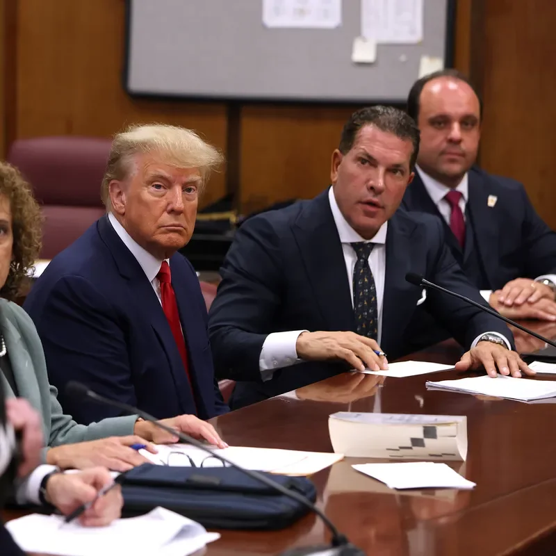 Former President Donald Trump sitting at a desk with security officer in foreground.