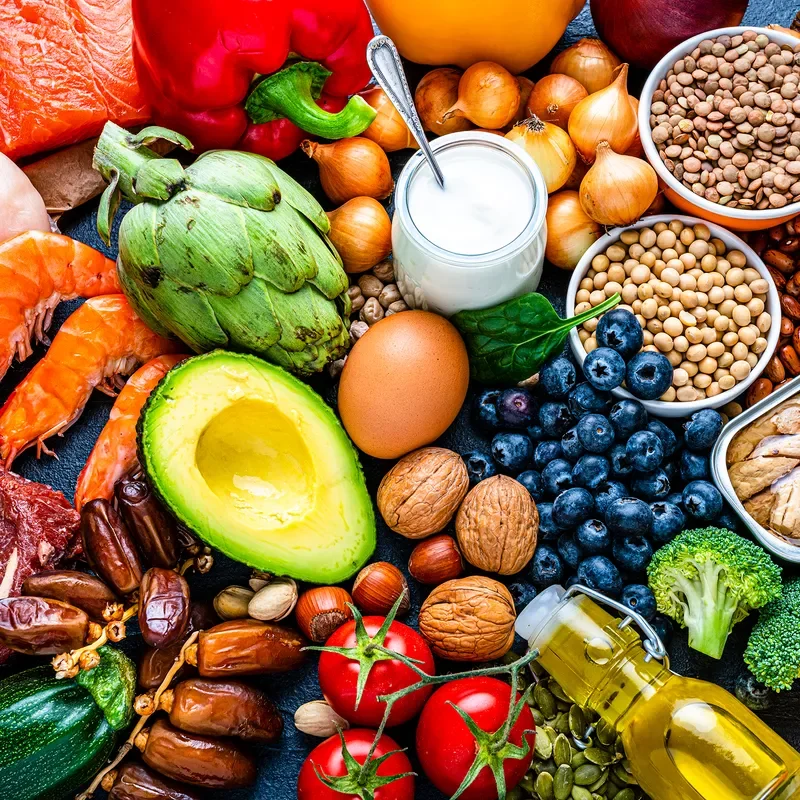 An overhead view of assorted foods including blueberries, avocado, tomatoes, meat, artichokes, and broccoli. 