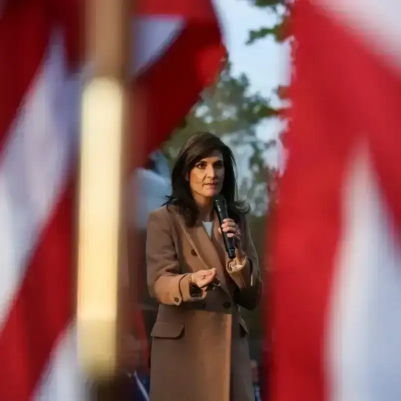 Republican presidential candidate Nikki Haley photographed in between two American flags. Haley wears a brown coat, holds a microphone in her left hand, and is gesturing with her right hand.