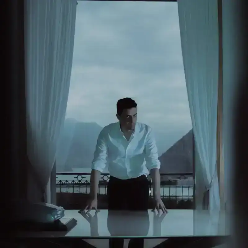 Hazim Nada posing behind a glass desk with both hands on the desk. He is wearing dark pants and a white button-up shirt. 