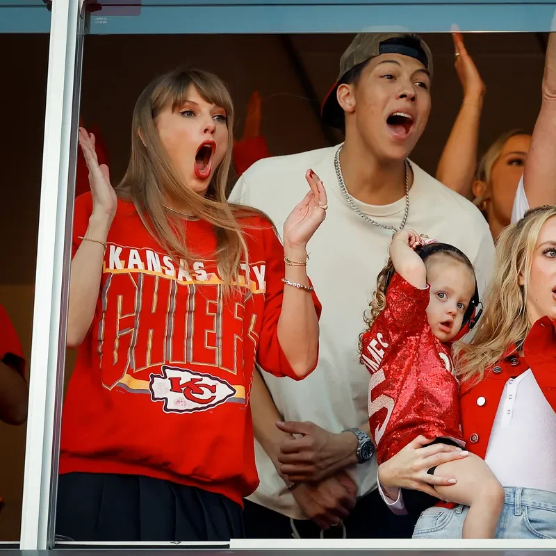 Taylor Swift wearing a Kansas City Chiefs sweatshirt and cheering with a crowd.