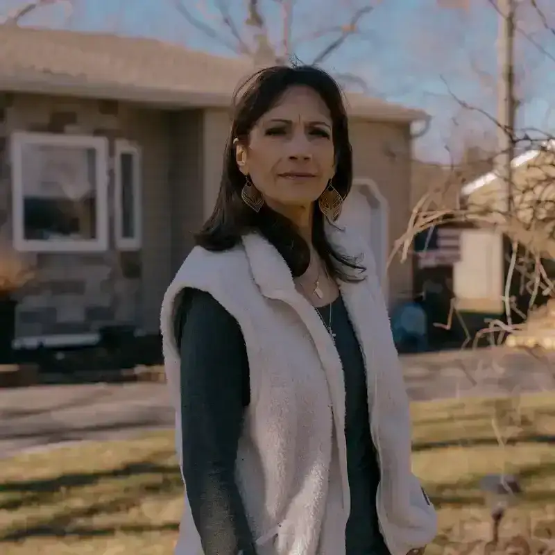 Luana Dunn standing outside of her home. Dunn wears blue jeans, a long-sleeve gray T-shirt, and a white vest. Her hair is dark brown and shoulder-length. 