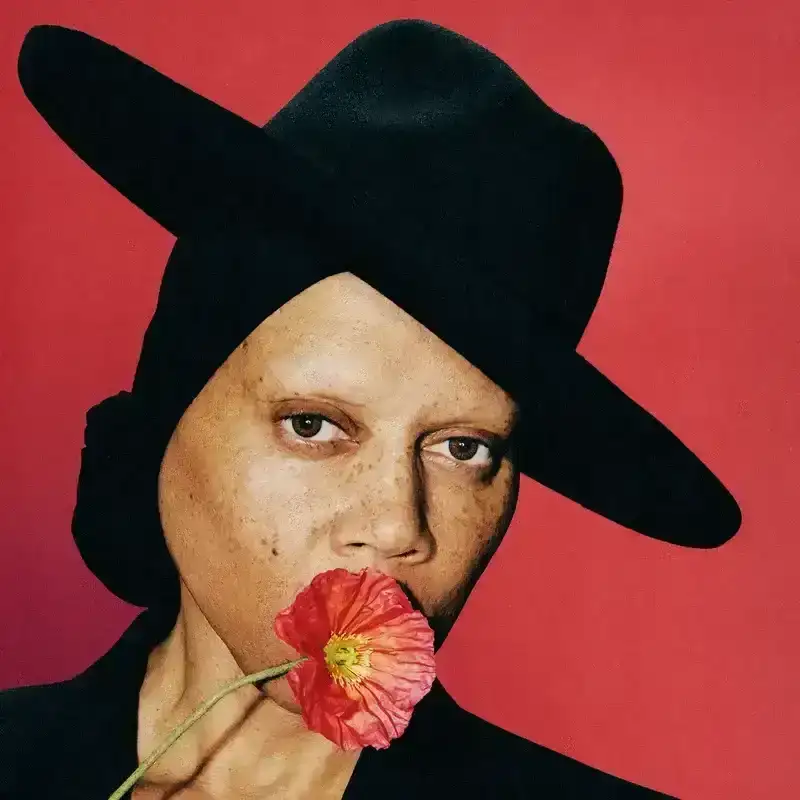 RuPaul wearing a black hat and black jacket, holding up a red flower in front of his mouth. 