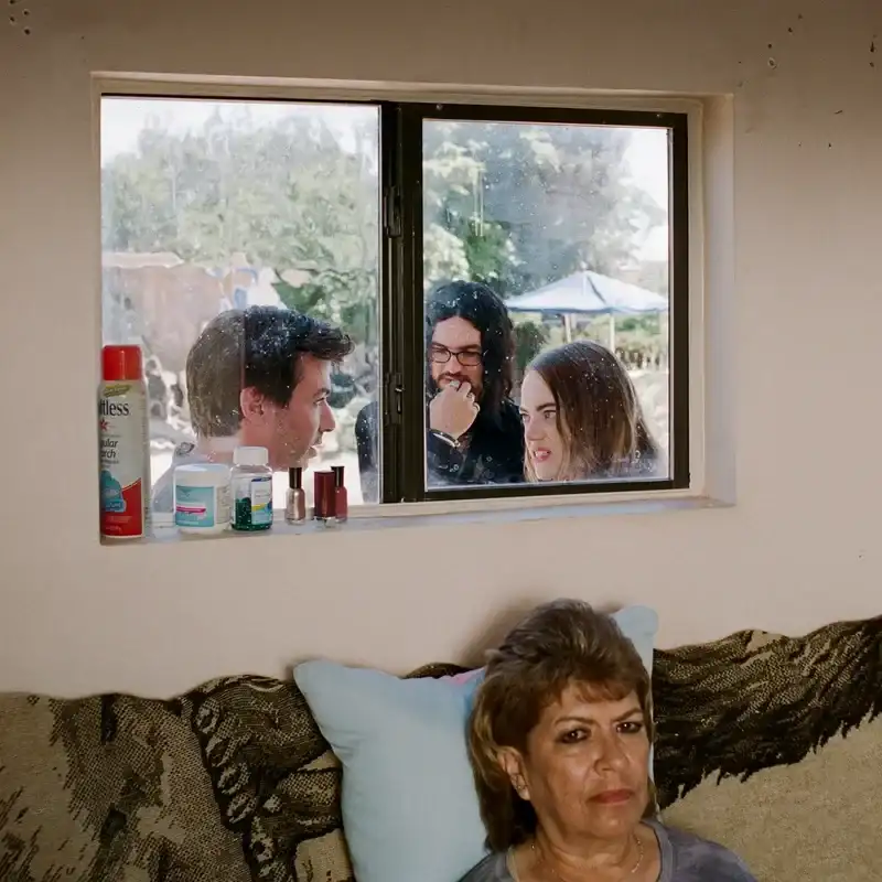 Nathan Fielder, Benny Safdie, Emma Stone, and Debbie Salazar in %22The Curse.%22 Fielder, Safdie, and Stone speak to one another outside and are seen through a window. Salazar is sitting inside a home on a couch. 