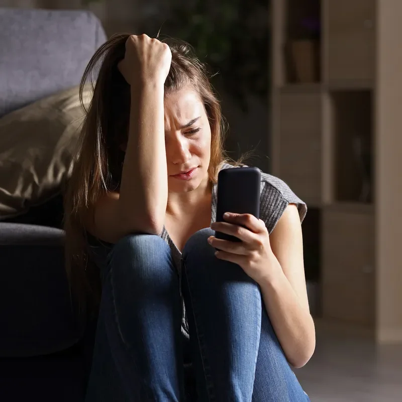 A sad teen checking phone sitting on the floor.